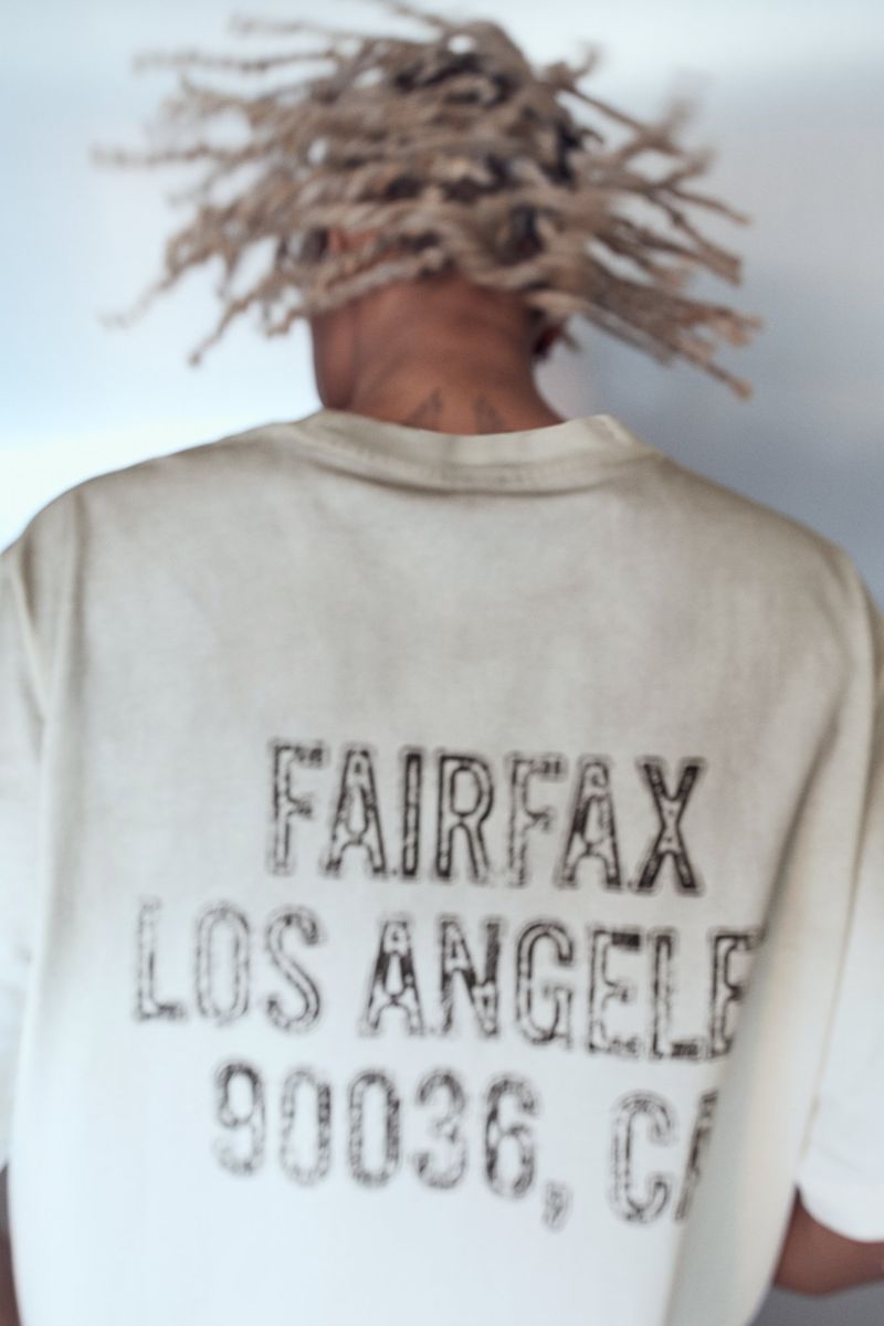 Person with blonde dreadlocks wearing a cream t-shirt with black Fairfax, Los Angeles, and 90036 CA text print viewed from behind.