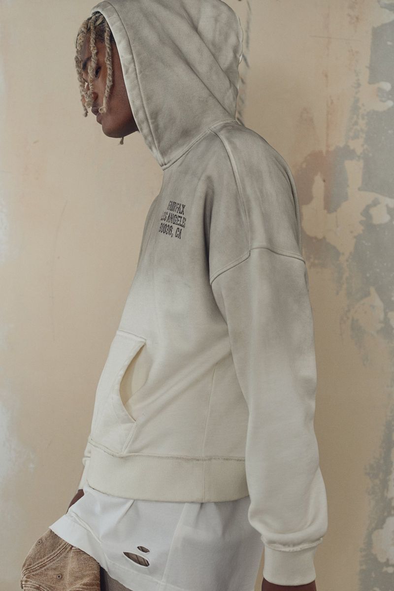 Fashion shot of a slender person wearing a gray and white hoodie with text, paired with white shorts and tan, distressed pants. 
