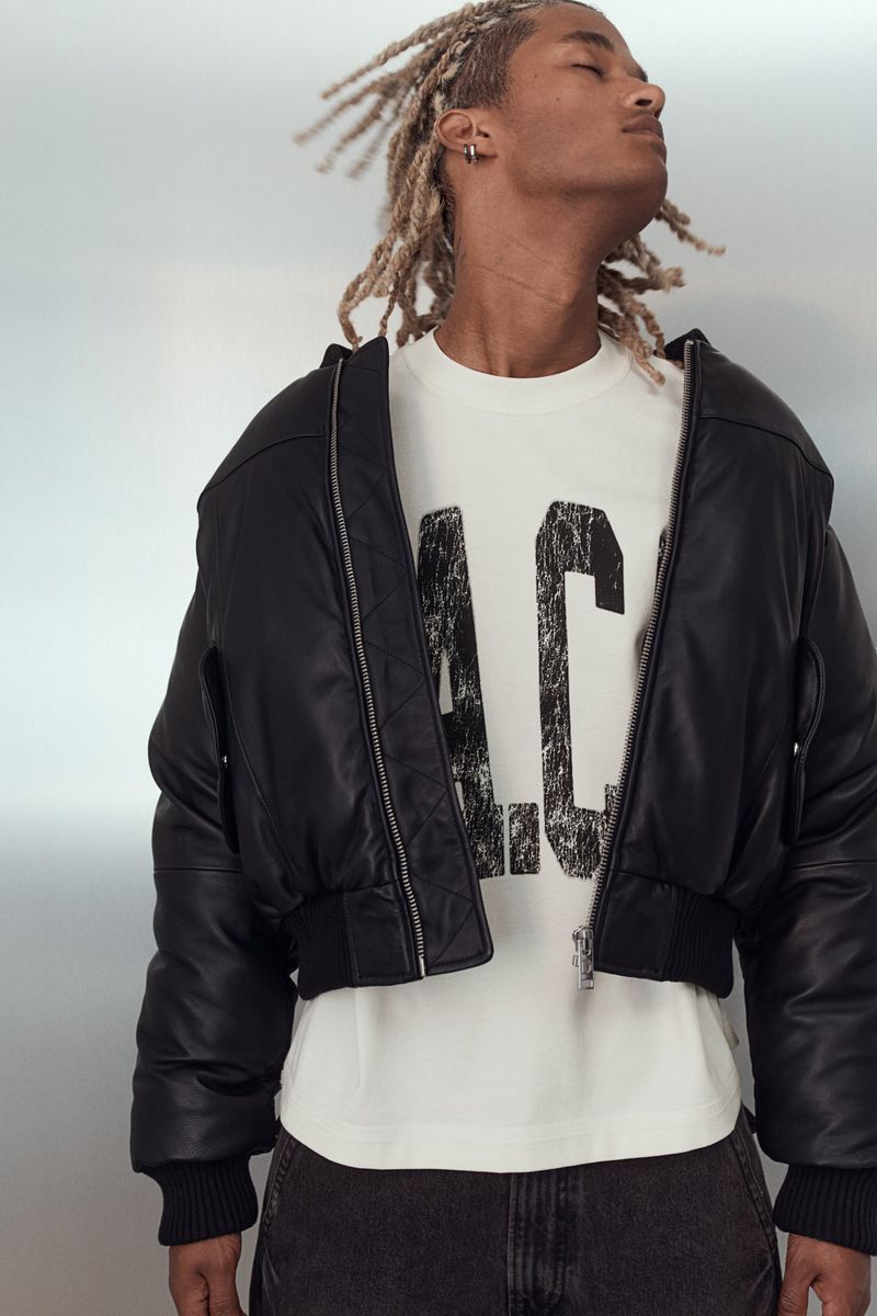 Close-up of a stylish adult male with blonde dreadlocks wearing a black bomber jacket and printed t-shirt against a grey background.