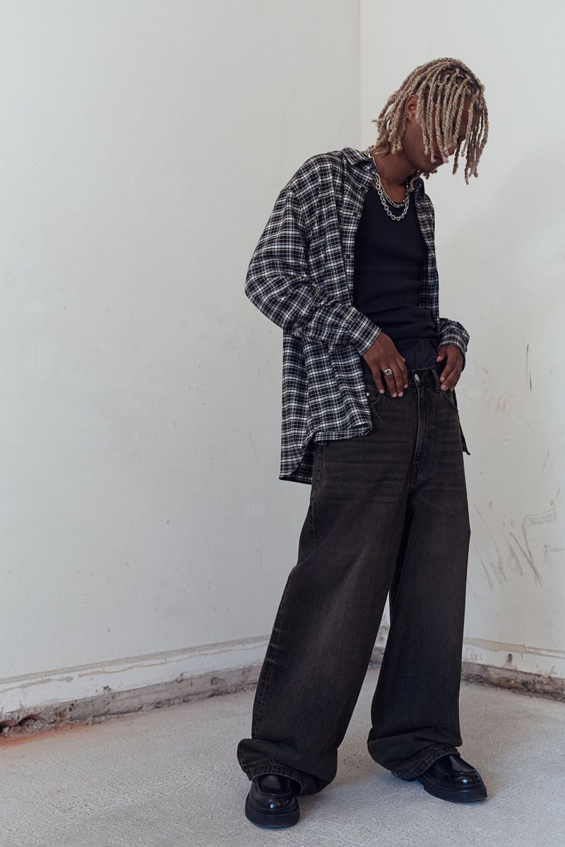 A young adult with dreadlocks wearing an unbuttoned black and white flannel shirt, black t-shirt, and wide black pants stands indoors.