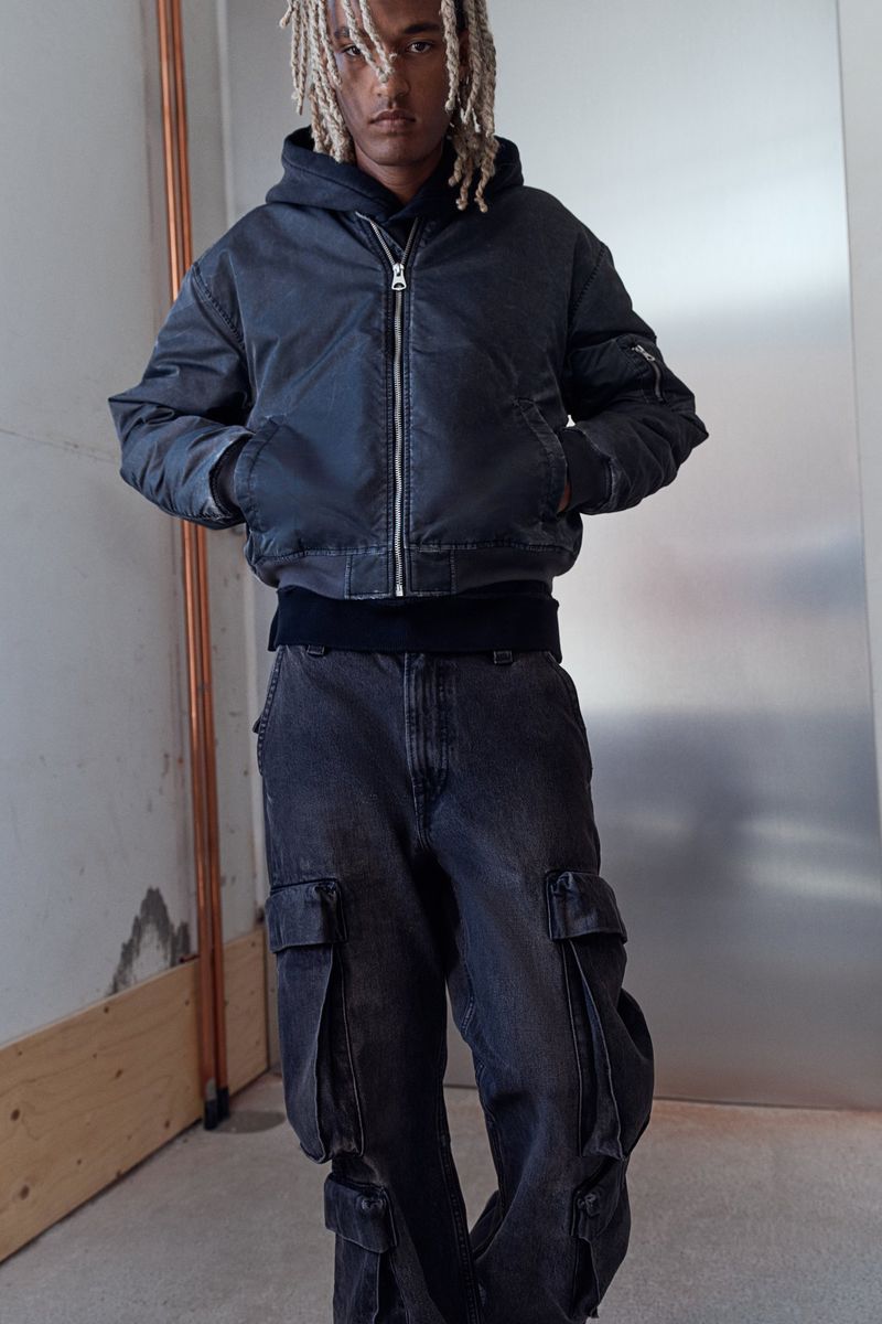 Young man with blond dreadlocks poses in a bomber jacket and baggy jeans against a neutral background indoors in a fashion shot.