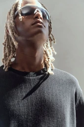 Close-up portrait of a man with blonde dreadlocks wearing dark sunglasses and a dark textured sweater against a light background.