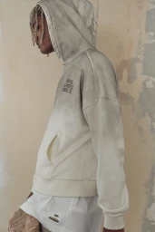 Model wearing a distressed cream hoodie with a hood up against a mottled neutral wall.