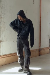 Model wearing a black hooded jacket and dark cargo pants posing against a textured wall interior.