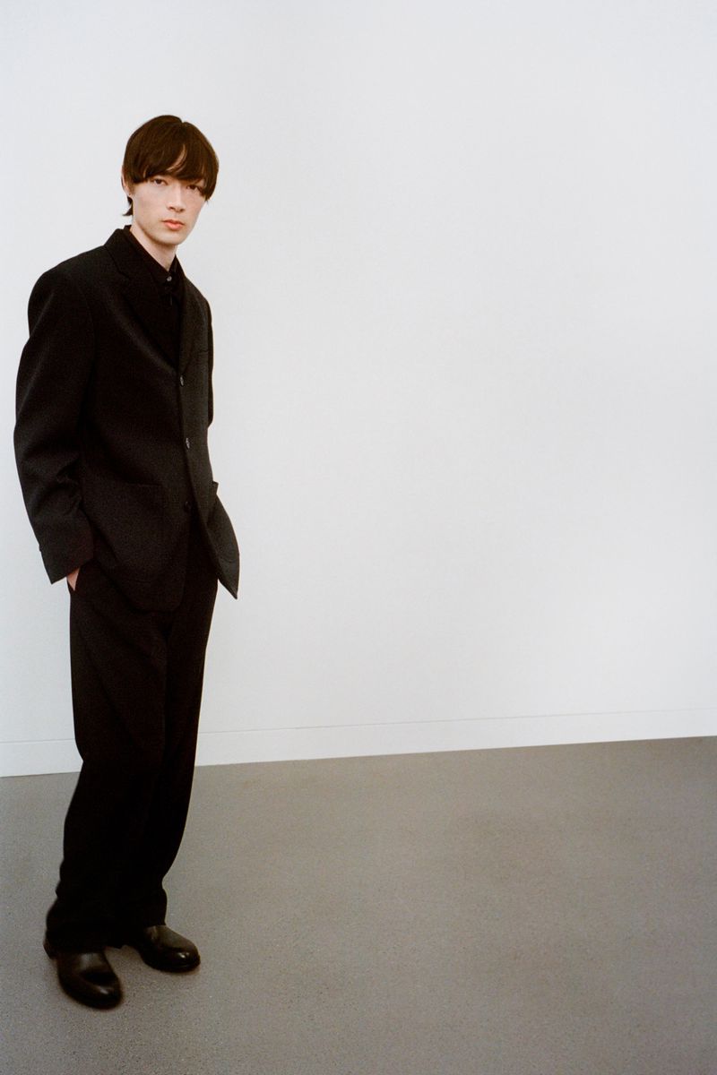 A young adult man stands in a black suit, black shirt, and black shoes, posed against a bright white wall and a light gray floor.