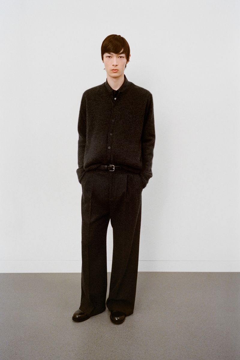 Young man in a dark button-down shirt, dark wide-leg trousers, and black dress shoes poses against a plain white wall in a studio.