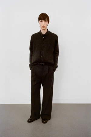 Young man in a dark button-down shirt, dark wide-leg trousers, and black dress shoes poses against a plain white wall in a studio.