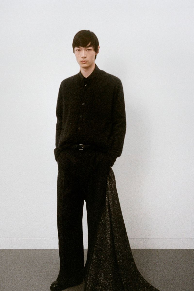 Portrait of a male fashion model in black attire posing in a photo studio with a neutral backdrop and soft lighting in a full-body shot.