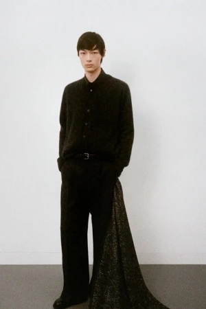 Portrait of a male fashion model in black attire posing in a photo studio with a neutral backdrop and soft lighting in a full-body shot.