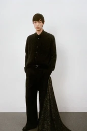 A male model wearing a dark knit top and trousers against a plain light wall background.
