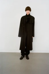 Model wearing a dark double-breasted trench coat and black trousers standing against a plain white studio backdrop.