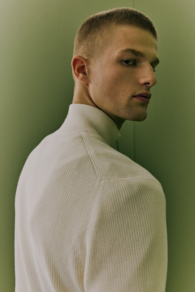 Close-up studio shot of a young adult male with short hair wearing a cream-colored ribbed turtleneck sweater against a light green backdrop.