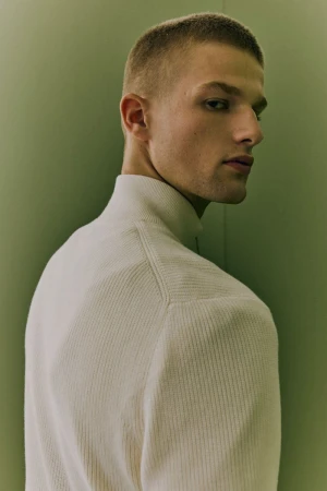 Close-up studio shot of a young adult male with short hair wearing a cream-colored ribbed turtleneck sweater against a light green backdrop.