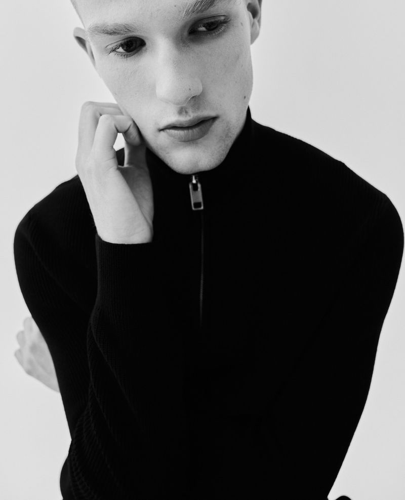 Black and white portrait of a young adult male in a black turtleneck sweater, facing forward with a hand gently touching his face.