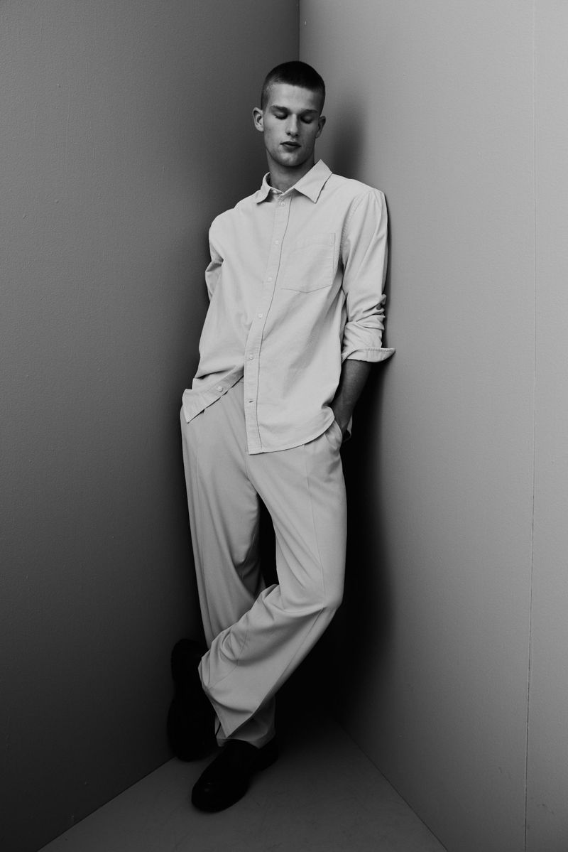 Monochrome portrait of a male model leaning against a corner wall wearing a button-down shirt and trousers for H&M.