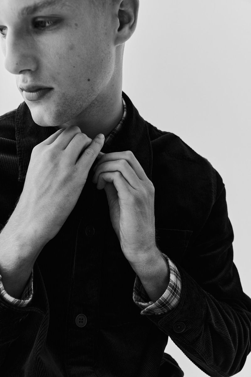 Monochrome portrait of a young adult male buttoning his corduroy jacket, with a plaid shirt collar visible, against a bright background.