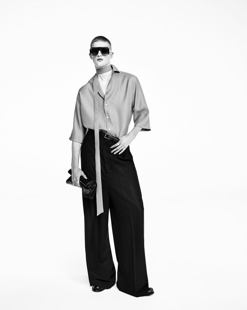 Black and white studio shot of a male model with dark sunglasses, button-down shirt, wide leg pants, holding a clutch purse.