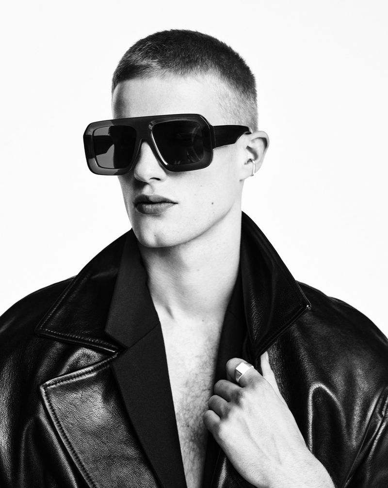 Black and white portrait of a young adult man with a buzzcut wearing oversized black sunglasses and a leather jacket in a photo studio.