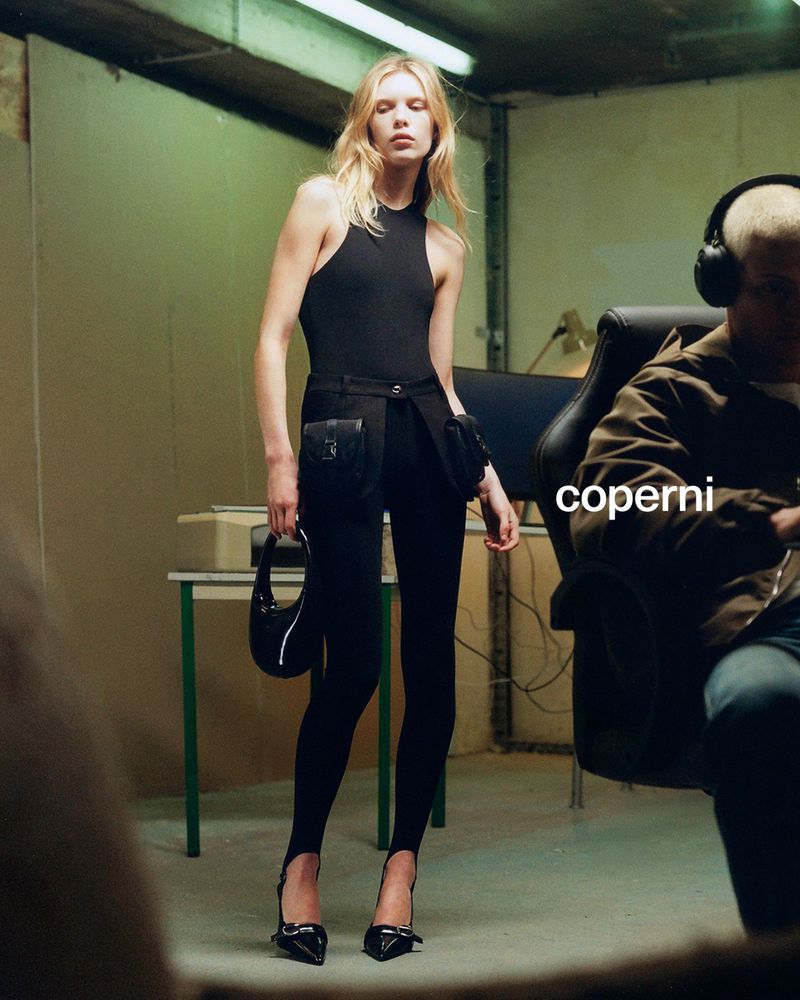 A blonde model poses in a black outfit holding a black purse in front of a seated person in a studio for a fashion editorial.