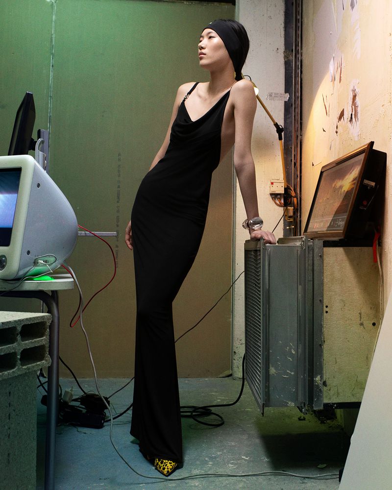 An Asian woman poses in a black dress in an industrial setting with a vintage computer and TV, exuding a retro-futuristic vibe.