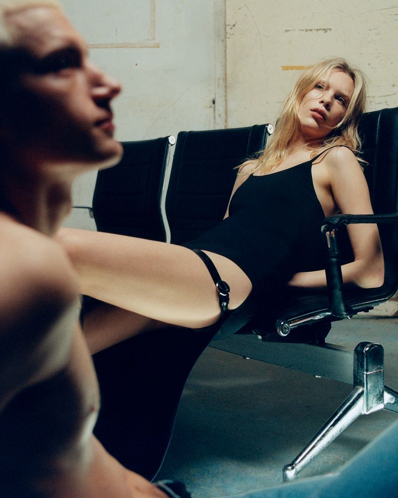 Blonde woman in black bodysuit reclines on black airport seating next to a seated fair-skinned male in a dimly lit indoor setting.