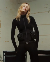 Model in black zip-up top with utility pouches posing against a neutral concrete background for Coperni