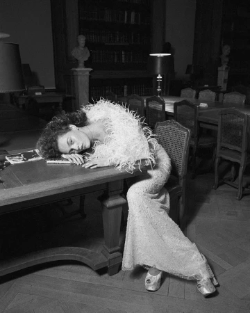 Woman in elegant feathered top and dress rests on a library table with books, in a black-and-white fashion shot, evoking timeless style.