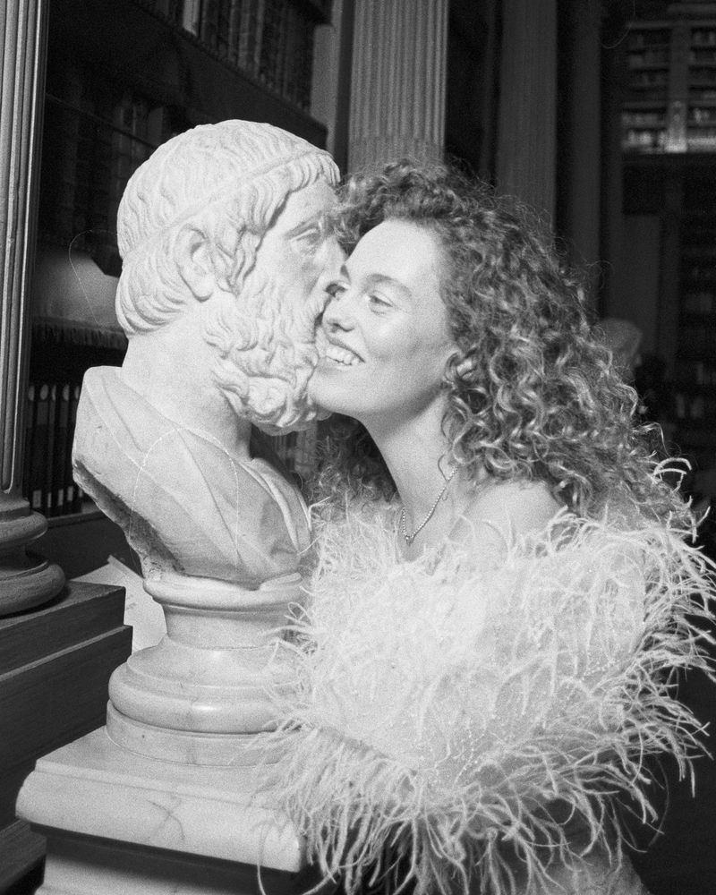 Monochrome image of a smiling woman in a feathered top affectionately touching her face to a marble bust in a library setting.