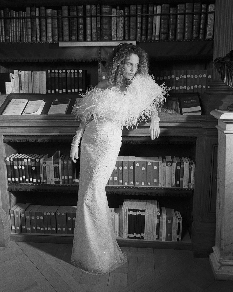 Woman in an embellished gown with feathered detail poses in front of a bookshelf filled with books in a black and white shot.