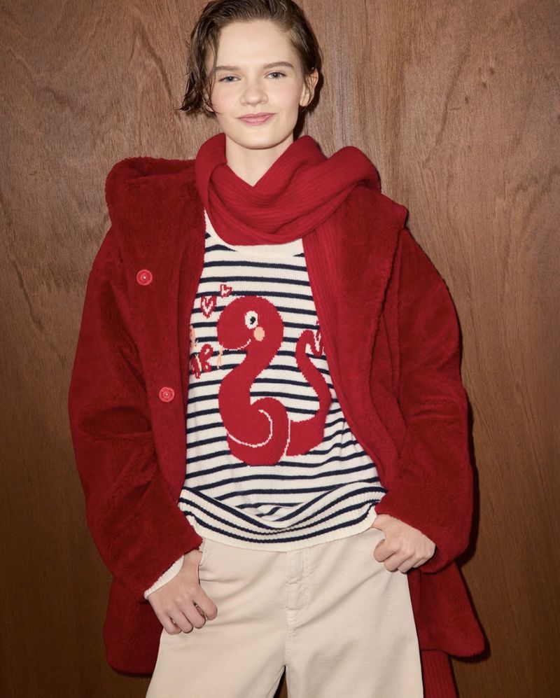 Teen girl poses indoors wearing a striped sweater with a red dinosaur, red coat, and light-colored pants in a fashion setting.