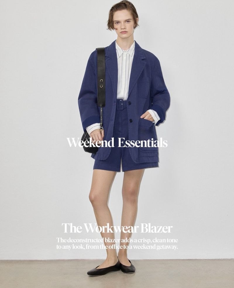 A woman models a navy blue blazer and shorts set with a white striped shirt and black bag, standing against a white backdrop in a studio.