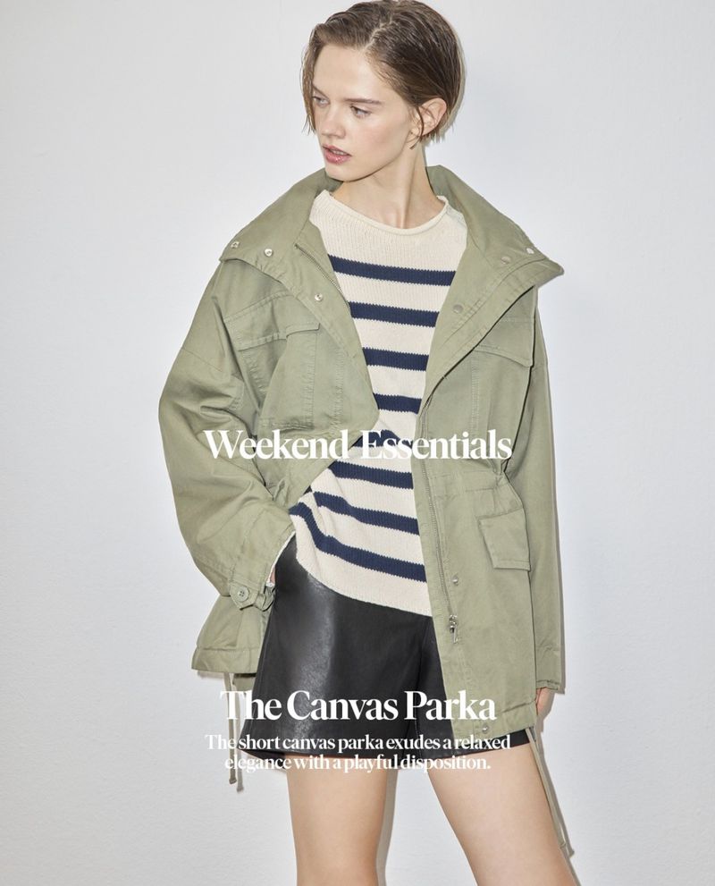 Woman in an olive canvas parka, white and navy striped sweater, and black shorts poses in front of a white studio backdrop.