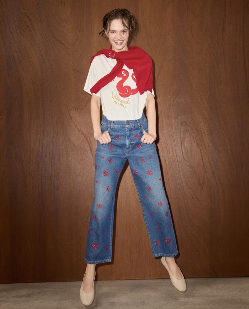 Smiling woman stands in blue floral-embroidered jeans, a white graphic tee, and red sweater in front of a wood paneled wall.