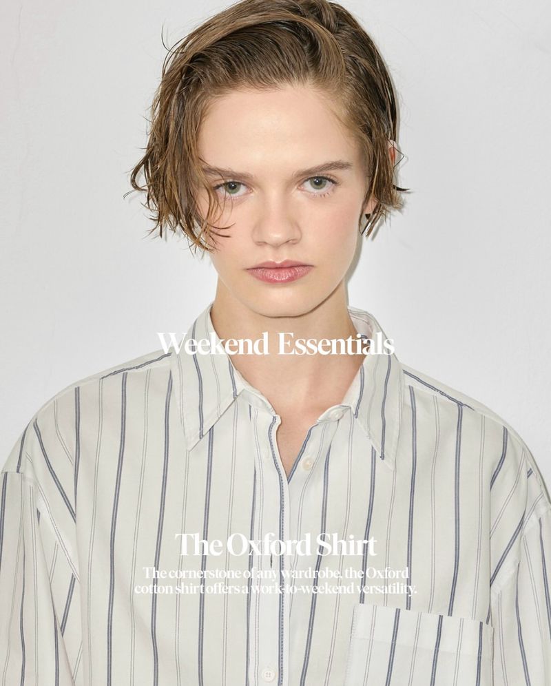 Close-up studio portrait of a young adult woman wearing a striped oxford shirt against a white backdrop, showcasing a fashion-forward aesthetic.