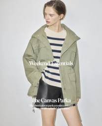 Model wearing an olive green canvas parka over a striped sweater and dark shorts against a white backdrop.
