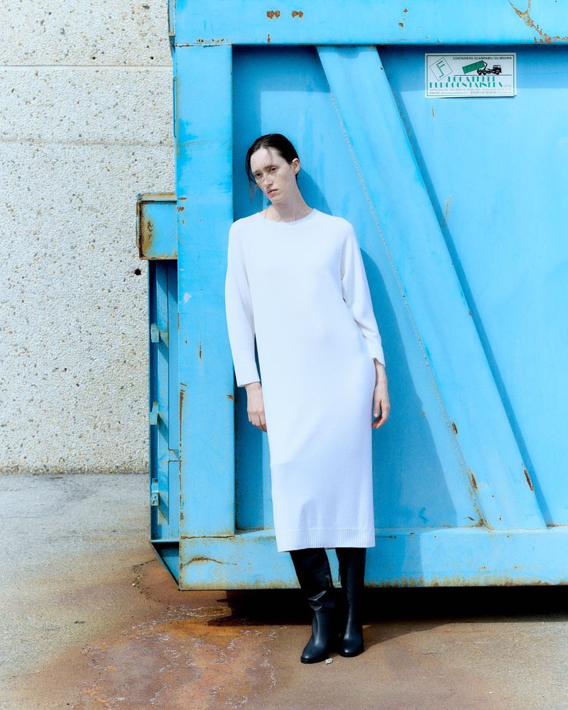 Adult woman in white dress stands next to blue dumpster.  She wears black boots in an outdoor setting next to a textured wall.