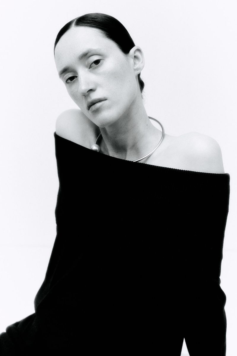 Elegant portrait of a woman with slicked-back hair and a dark off-the-shoulder dress against a bright white backdrop in a studio setting.