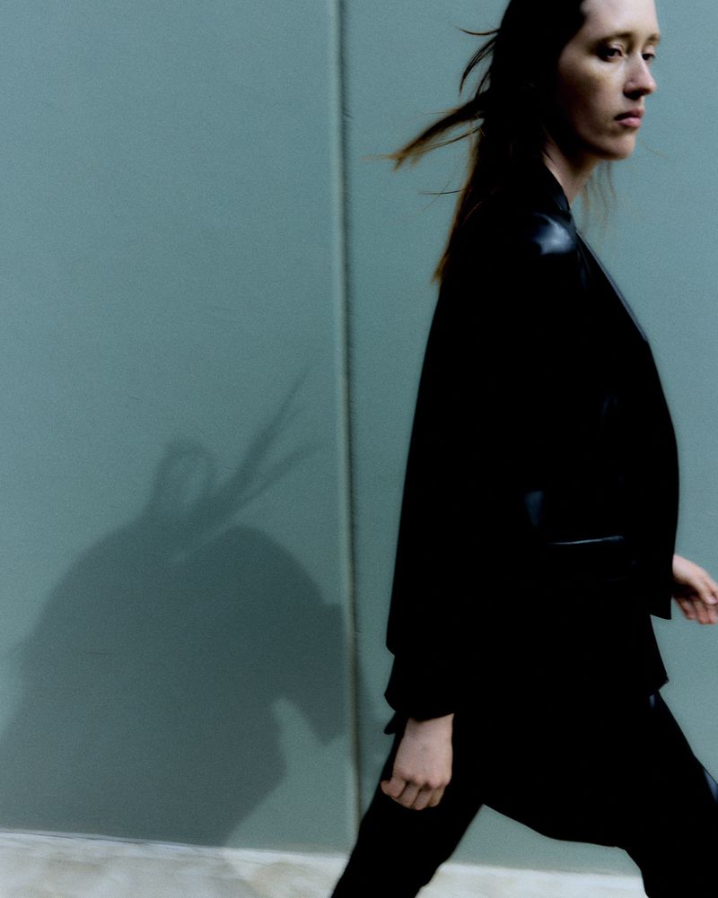 Fashion shot of a woman in a black suit walking against a grey-green background. Her hair is blowing, and a shadow is on the wall.