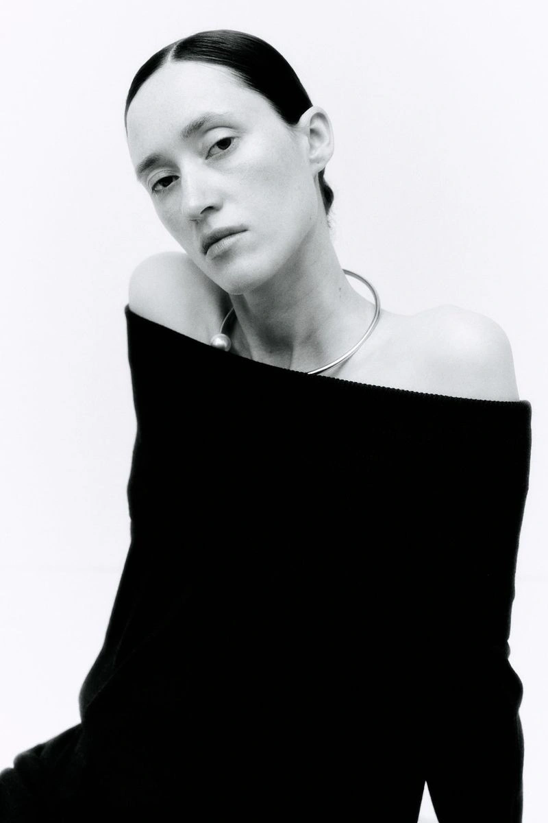 Model wearing an off-the-shoulder black top and silver collar necklace against a clean white background