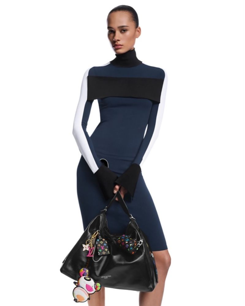 Model in color-blocked turtleneck dress holding a black leather handbag against a plain white background.