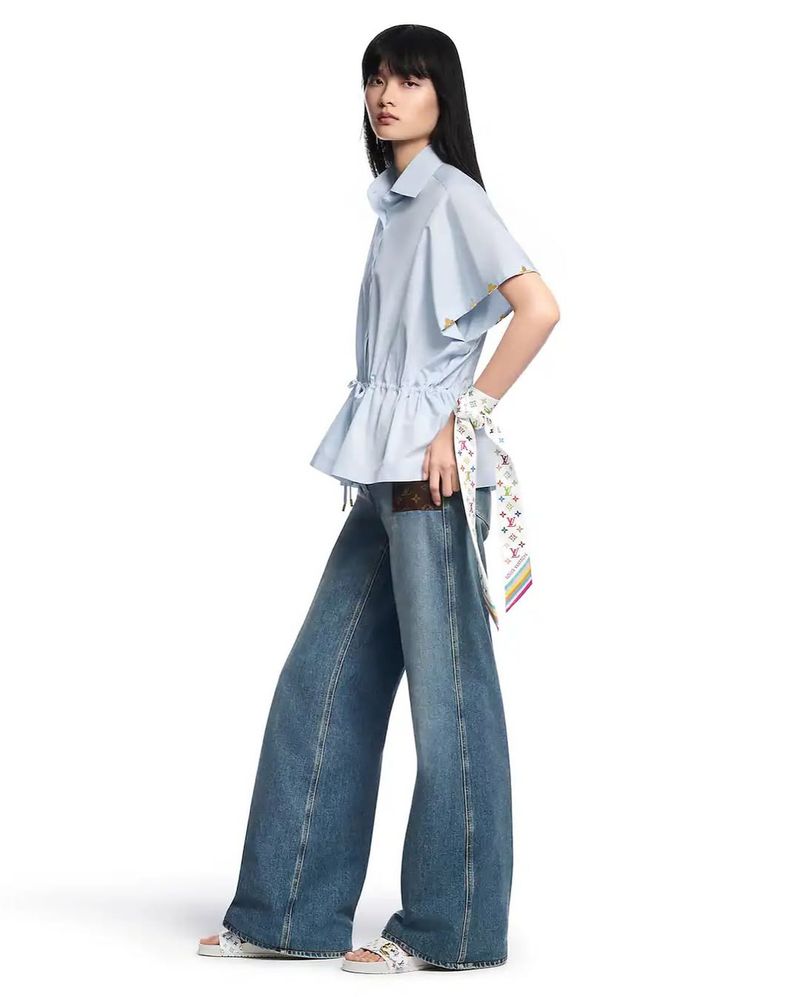 Fashion shot of an Asian woman in blue blouse and wide-leg jeans, holding a purse and wearing patterned sandals against white backdrop.