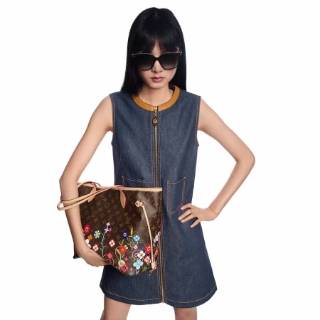 Woman in a denim dress holds a brown Louis Vuitton handbag with floral embroidery against a white background in a studio setting.