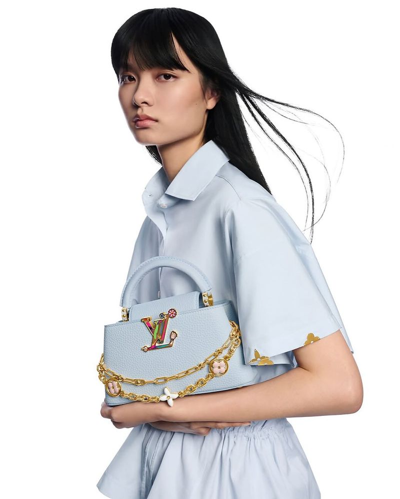 Asian woman models a light blue Louis Vuitton Capucines handbag with a gold chain against a white backdrop in a fashion studio shot.