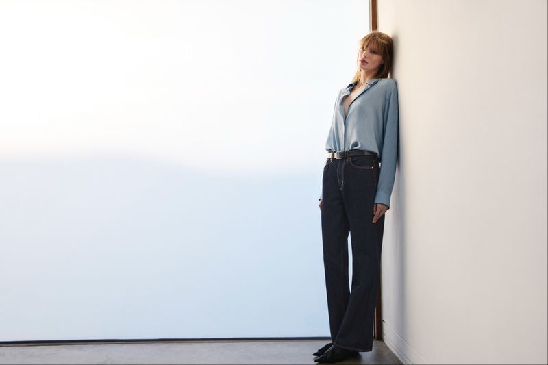 A woman in a blue top and dark jeans leans against a white wall in a fashion-style photograph with soft lighting and indoor setting.