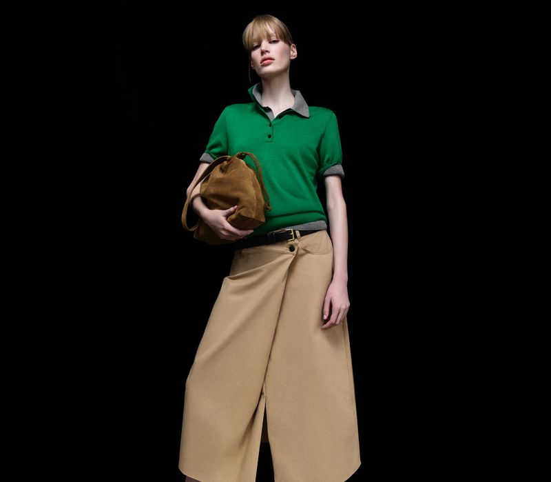 Fashionable blonde woman in a green collared shirt and khaki skirt holding a suede purse against a black backdrop in a studio.