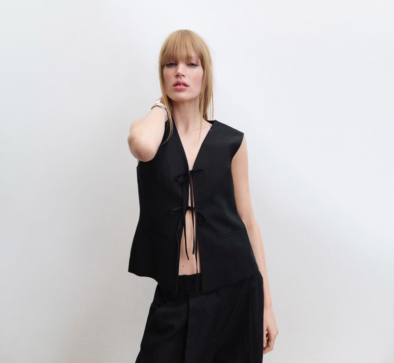 Model with long blonde hair and bangs wearing a black tie-front vest against a plain white background