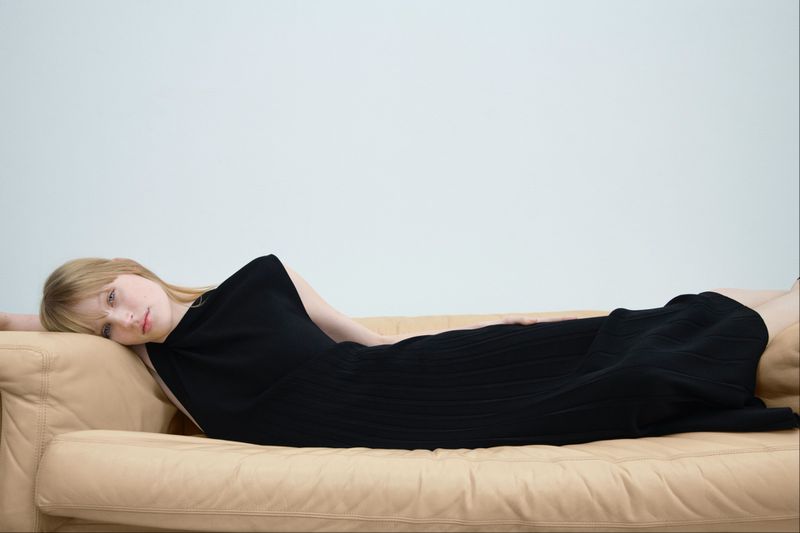 Blonde woman reclining on a beige leather sofa wearing a black sleeveless dress against a plain neutral background.