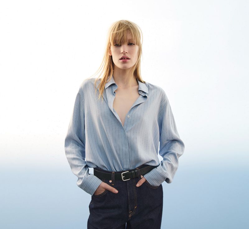 Fashion shot of a blonde adult woman in a light blue striped shirt, dark jeans, and black belt, posing outdoors in diffused lighting.