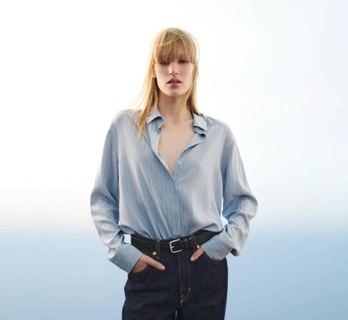 Fashion shot of a blonde adult woman in a light blue striped shirt, dark jeans, and black belt, posing outdoors in diffused lighting.