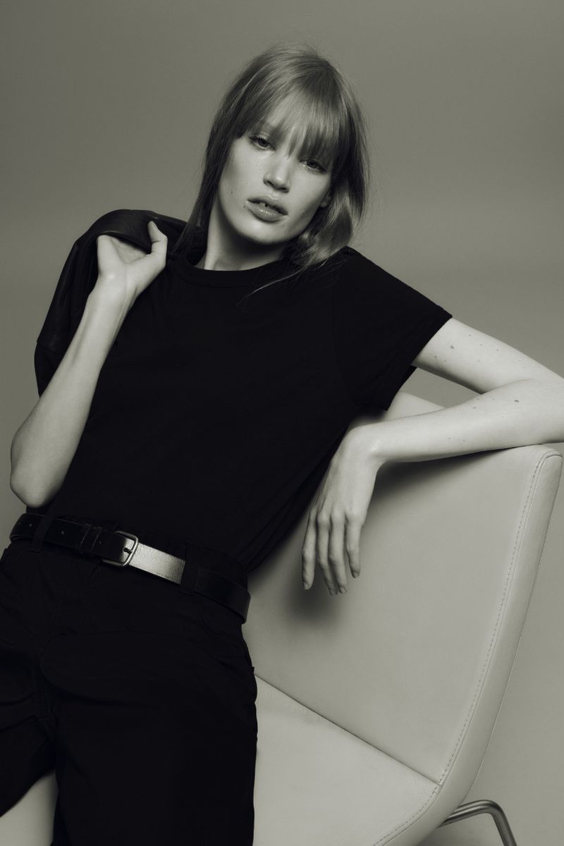 Blonde woman in black t-shirt and pants poses in studio against grey backdrop with jacket slung over shoulder while sitting in chair.
